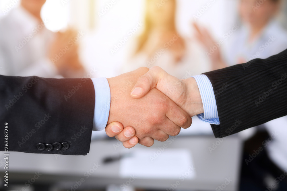 Business people shaking hands at meeting while theirs colleagues clapping and applauding. Group of unknown businessmen and women in modern white office. Success teamwork, partnership and handshake