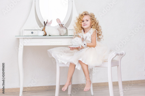 Little beautiful and cute girl child in a fashionable festive dress and with animals rabbits in the mirror in a white interior