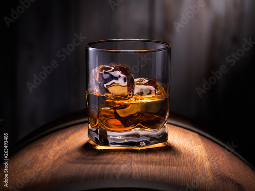 Fotografia Glass of whiskey with ice cubes on the wooden barrel with wooden background
