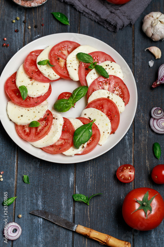 Italian caprese salad in white plate with sliced tomatoes. Delicious italian caprese salad with ripe tomatoes, fresh basil and mozzarella cheese on dark wooden rustic background.