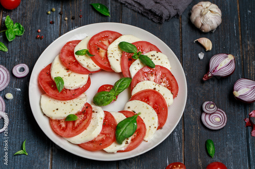 Italian caprese salad in white plate with sliced tomatoes. Delicious italian caprese salad with ripe tomatoes, fresh basil and mozzarella cheese on dark wooden rustic background.