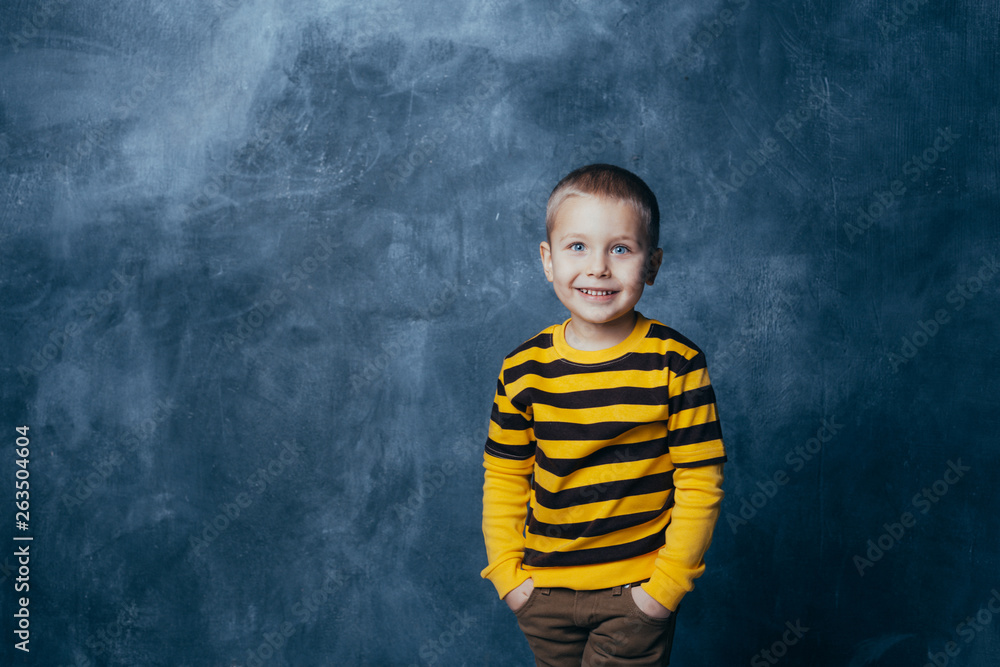 A little boy poses in front of a gray-blue concrete wall. Portrait of a smiling child dressed in a black and yellow striped sweater and brown pants.