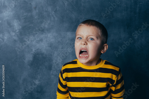  Portrait of a screaming child with open mouth, A little boy poses in front of a gray-blue concrete wall.