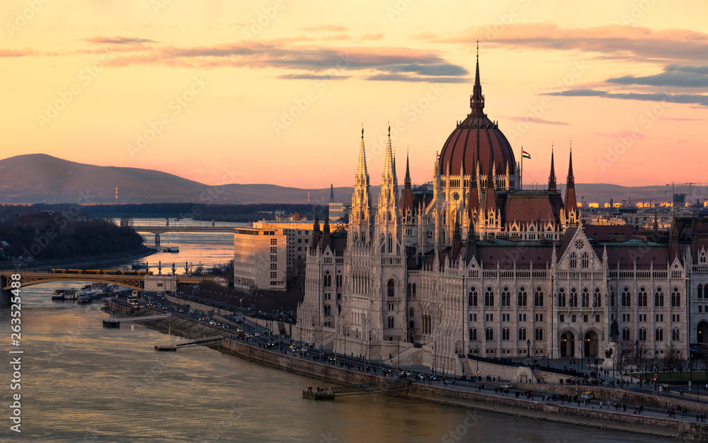 Cityscape of Budapest with bright parliament illuminated by last sunshine before sundown and Danube river with bridge. Pink and purple colors of sky reflecting in water during sunset.