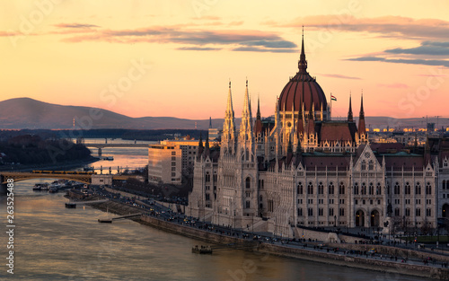 Cityscape of Budapest with bright parliament illuminated by last sunshine before sundown and Danube river with bridge. Pink and purple colors of sky reflecting in water during sunset. © Martin
