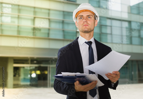 Smiling engineer in suit and hat with folder is exploring documents with project photo