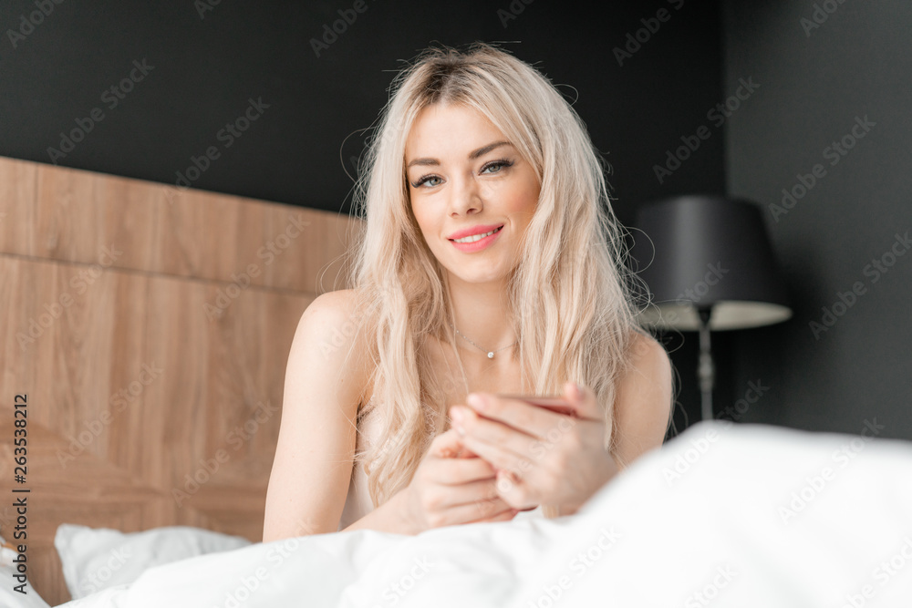 Use the phone. Checks email and social media messages. Young woman wake up, sits on comfortable bed. Morning in hotel room. Use the phone. Checks email and social media messages. Young woman wake up, sits on comfortable bed. Morning in hotel room.
