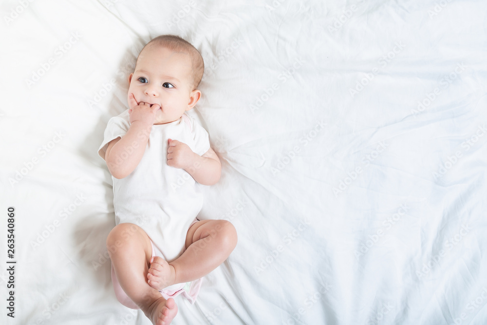 The baby teething Close up portrait of little baby girl in white lying on a white bed. The baby teething Close up portrait of little baby girl in white lying on a white bed.