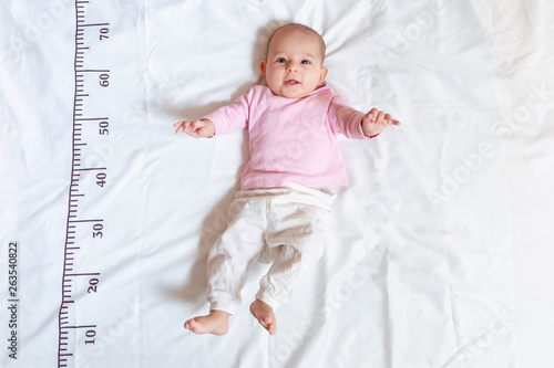 A four month happy baby in pink white clothes lying on a bed on which a measuring ruler for growth is drawn.