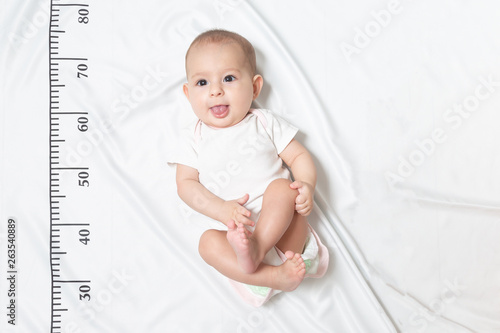 A five month baby in white clothes lying on a bed on which a measuring ruler for growth is drawn. shows the tongue from her mouth and teased