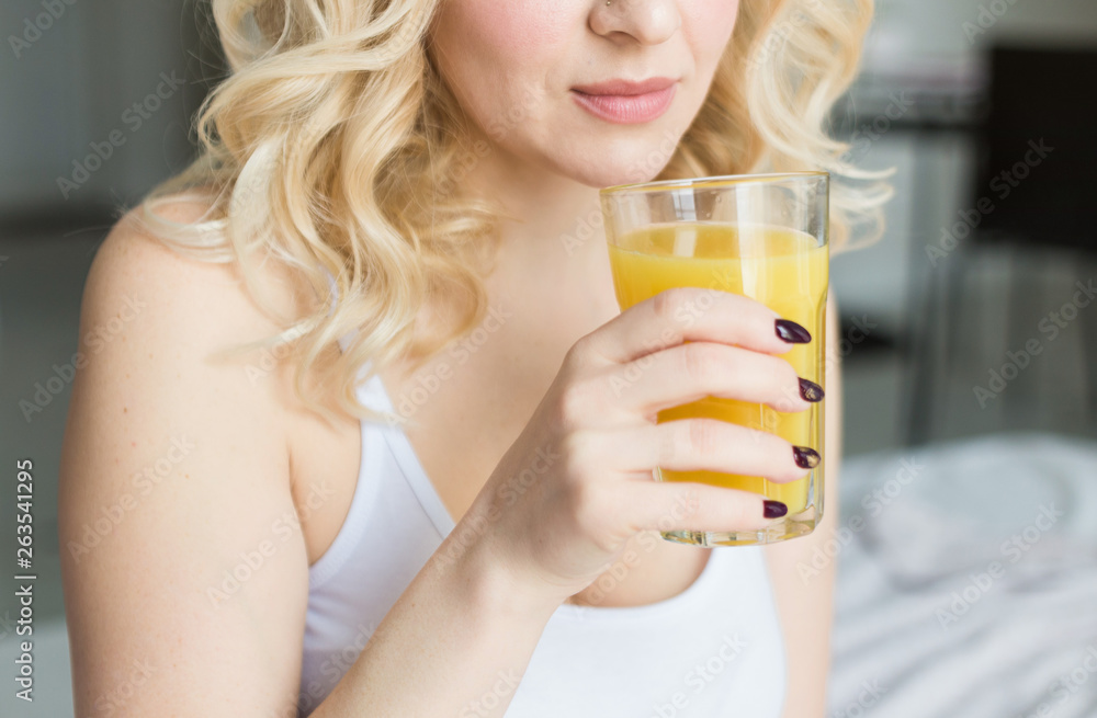 Attractive blonde woman sits on the edge of the bed at home and holds a stocker with orange juice in her hand. Morning mood and vigor Attractive blonde woman sits on the edge of the bed at home and holds a stocker with orange juice in her hand. Morning mood and vigor