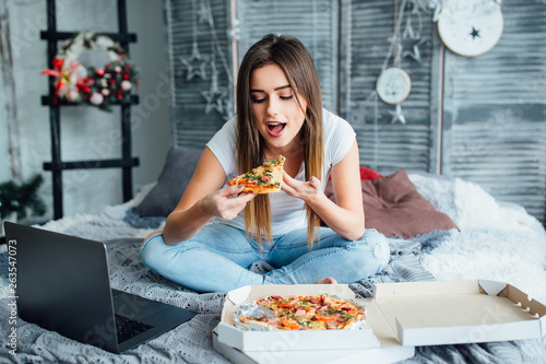 Cheerful beautiful female ordered pizza home. Woman eating in bed. Tasty Italian snacks. photo