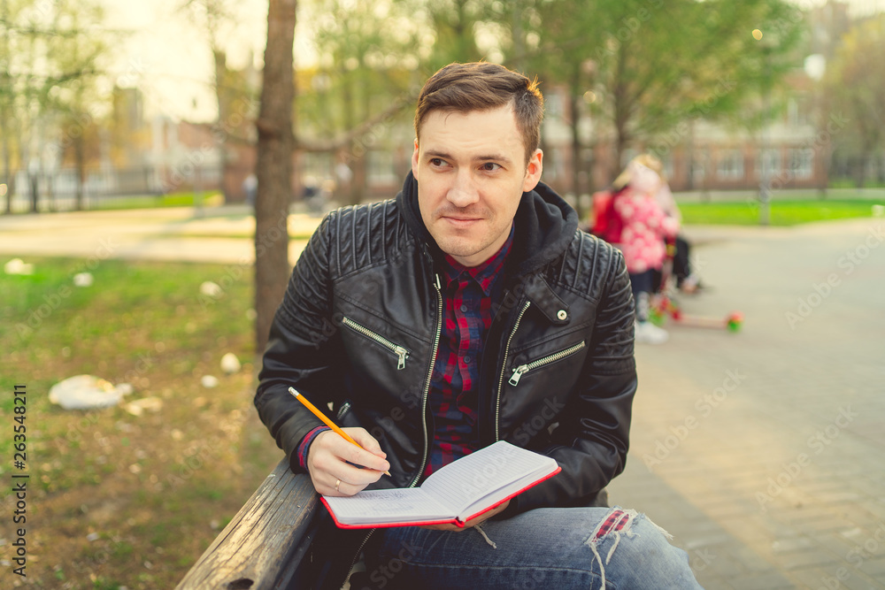 Obraz premium Man with a notebook in his hands in the Park. Male, student is writing her ideas and thoughts into the notebook sitting on the bench in the park 