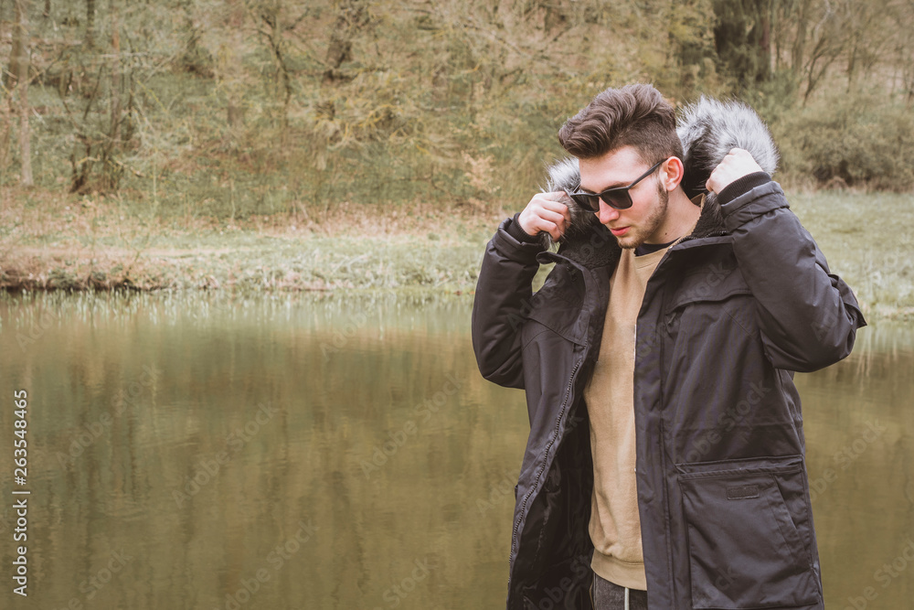 Portrait of a fashionable young guy wears a stylish furry jacket and posing near a pond. Portrait of a fashionable young guy wears a stylish furry jacket and posing near a pond.