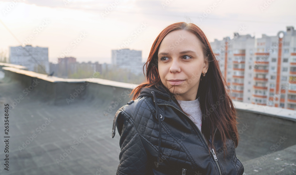 Woman in trendy jacket on roof edge. Portrait of young female in leather jacket, standing on the roof of at sunset