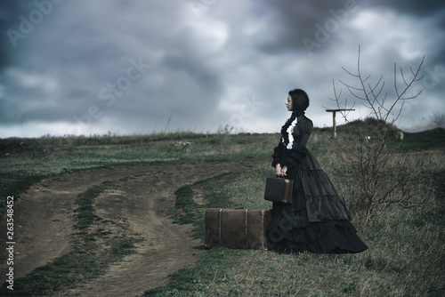Outdoors portrait of a victorian lady in black sitting alone on the road with her luggage. photo