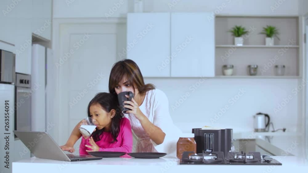Young mother and her daughter using a laptop computer while enjoying hot drinks on the morning in the kitchen at home Young mother and her daughter using a laptop computer while enjoying hot drinks on the morning in the kitchen at home