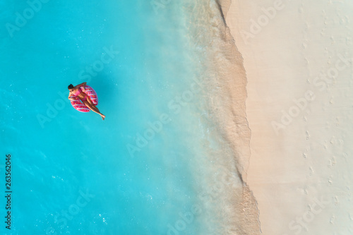 Aerial view of a young woman swimming with the donut swim ring in the clear blue sea with waves at sunset in summer. Tropical aerial landscape with girl, azure water, sandy beach. Top view. Travel