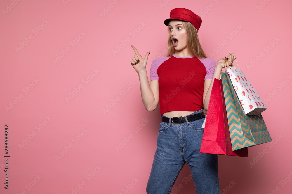 Young beautiful shocked girl in red hat with colorful shopping bags pointing up with finger, looking away Young beautiful shocked girl in red hat with colorful shopping bags pointing up with finger, looking away