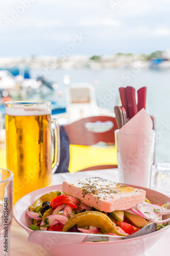 Bowl of fresh Greek salad with feta cheese, green pepper, tomato, cucumber, olives and red onion. Greek salad and beer mug on table against the sea bay background in Greece
