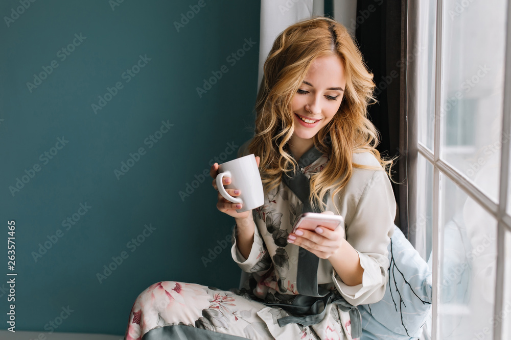 Pretty blonde girl sitting on window sill with cup of coffee, tea and smartphone in hands. She has long blonde wavy hair, smile and looking at her phone. Wearing beautiful silk pajama. - obrazy, fototapety, plakaty 