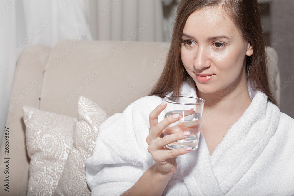 Young pretty european woman holding a glass and drink pure water sitting on the sofa. Cleansing the body of toxins. Healthy skin. Oily skin. Close up partrait of beautiful woman in bathrobe. Young pretty european woman holding a glass and drink pure water sitting on the sofa. Cleansing the body of toxins. Healthy skin. Oily skin. Close up partrait of beautiful woman in bathrobe.