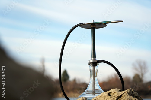 Fototapeta Naklejka Na Ścianę i Meble -  Blue wooden hookah with clear water with a dark hose stands in nature on the stone. Mouthpiece on a metal plate. Travel and hookah smoking concept. Blurred background with a cloudy sky.