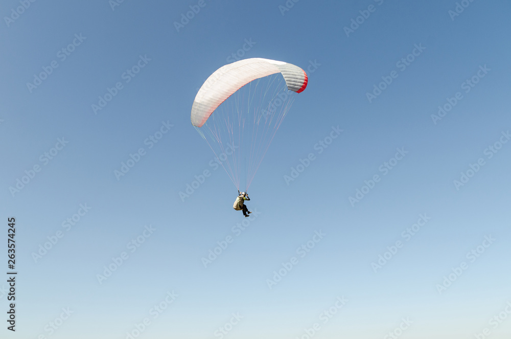 A man flies on a paraglider over green fields in the hot summer. Against the background of blue sky and rare clouds A man flies on a paraglider over green fields in the hot summer. Against the background of blue sky and rare clouds