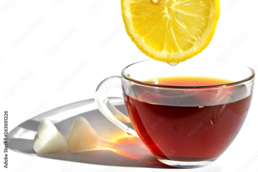 Glass cup of tea with long rainbow shadow in morning rays of sunlight, pieces of sugar in shape of heart and slice of sour eellow lemon with drop of refreshing juice on white background. Isolated Glass cup of tea with long rainbow shadow in morning rays of sunlight, pieces of sugar in shape of heart and slice of sour eellow lemon with drop of refreshing juice on white background. Isolated