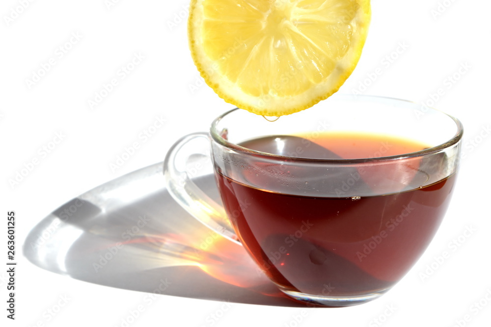 Yellow slice of bitter sour lemon with a drop of refreshing juice over transparent glass cup of hot black tea with long rainbow shadow in morning rays of sunlight on white background. Isolated Yellow slice of bitter sour lemon with a drop of refreshing juice over transparent glass cup of hot black tea with long rainbow shadow in morning rays of sunlight on white background. Isolated