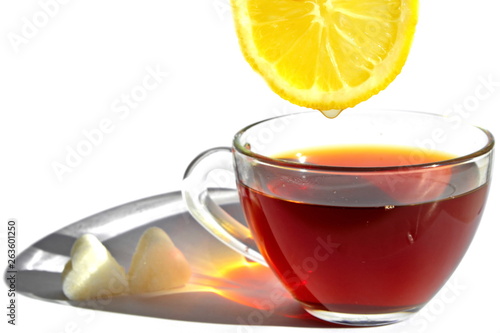 Yellow slice of sour lemon with drop of refreshing juice over glass cup of tea with long rainbow shadow in morning rays of sunlight and pieces of sugar in shape of heart on white background. Isolated