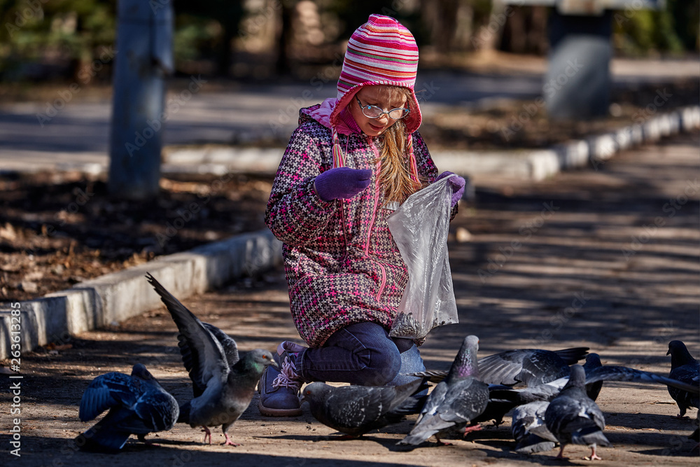 A girl child in a red jacket, hat and glasses feeds doves in the park with seeds. A girl walks along a park alley on a sunny spring day. A girl child in a red jacket, hat and glasses feeds doves in the park with seeds. A girl walks along a park alley on a sunny spring day.