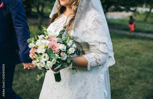 Wedding couple walking in the green park. Curvy bride in white lace dress and groom are holding hands. Overweight happy people. Love story outdoors. Beautiful bouquet.