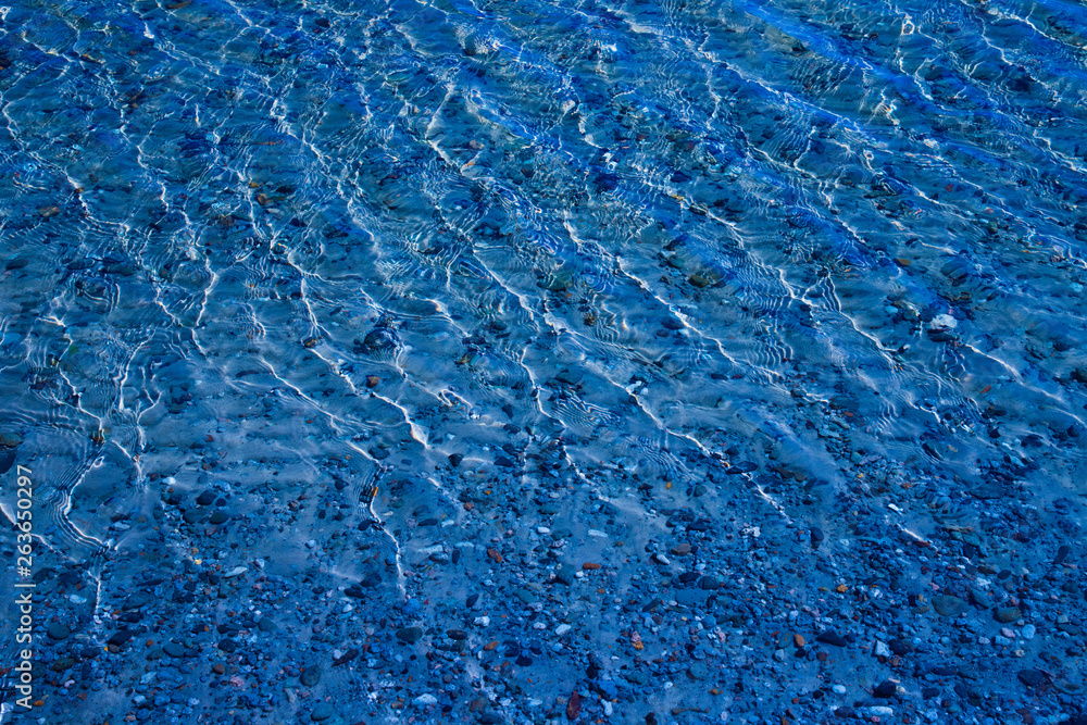 Blue water surface with ripples through which the sandy bottom with pebbles is visible Blue water surface with ripples through which the sandy bottom with pebbles is visible