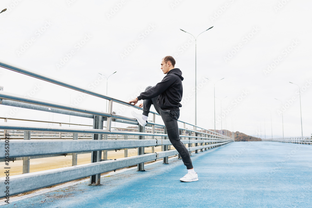 Side view - a handsome slender young male in a gray tracksuit kneads the joints and ligaments of the legs to a street workout standing on the bridge. Copyspace