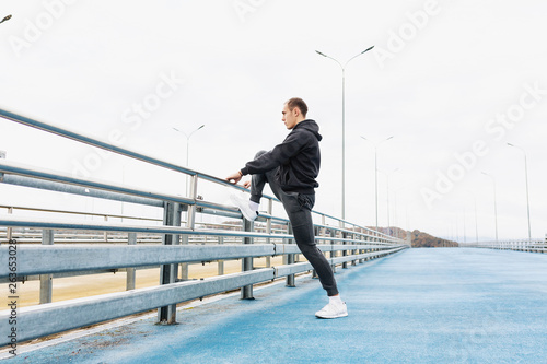 Side view - a handsome slender young male in a gray tracksuit kneads the joints and ligaments of the legs to a street workout standing on the bridge. Copyspace © Rithor