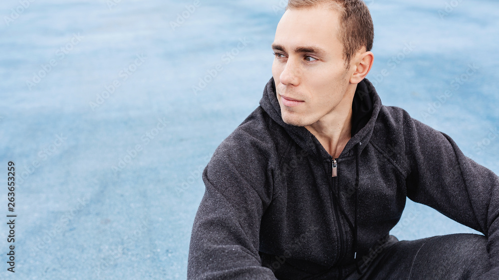 Young fit man in a gray sports suit sitting on the cover of the sports field resting after the morning run preparing for strength training. Healthy lifestyle and body concept. Copyspace Young fit man in a gray sports suit sitting on the cover of the sports field resting after the morning run preparing for strength training. Healthy lifestyle and body concept. Copyspace