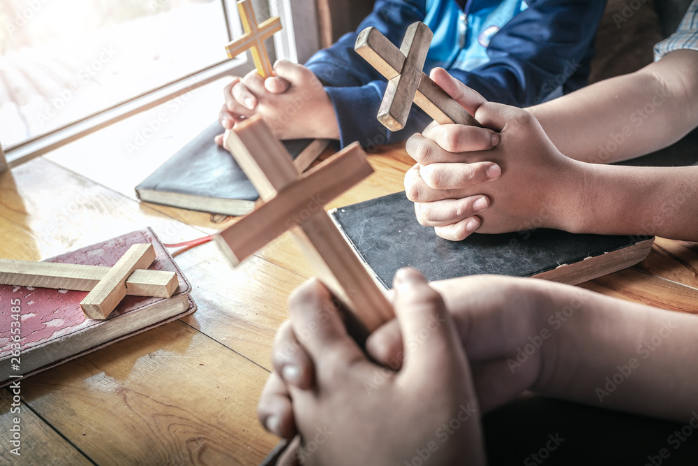Christian children group holding christian cross and praying together around wooden table with open bible page at home, prayer meeting concept.