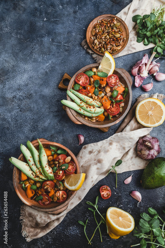 vegetarian salad with roasted pumpkin, chickpea, tomatoes, pumpkin and sunflower seeds, avocado, lemon and spices in bowls on blue background, copy space
