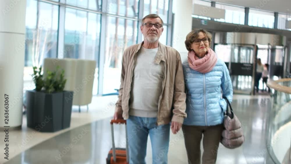 Mature woman and man walk through the airport terminal. The man has a suitcase in his hands. Romantic travel couple of pensioners Mature woman and man walk through the airport terminal. The man has a suitcase in his hands. Romantic travel couple of pensioners