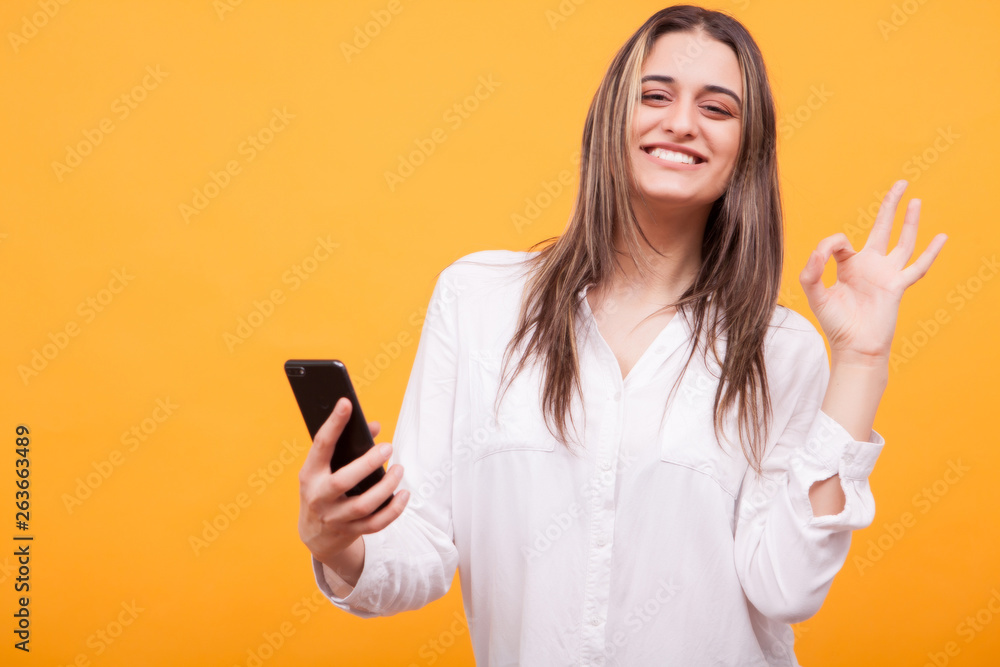Beautiful girl in white shirt holding phone and showing ok sign over yellow background
