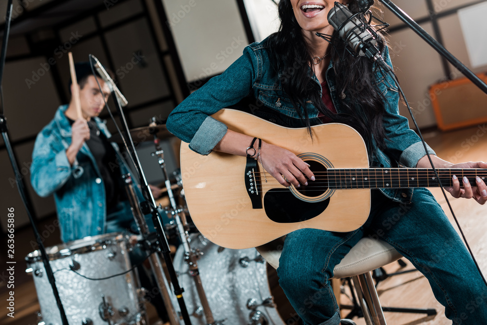 partial view of musician playing guitar and singing while mixed race man playing drums in recording studio partial view of musician playing guitar and singing while mixed race man playing drums in recording studio