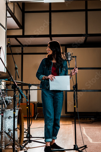 beautiful singer standing near microphone and holding paper with text in recording studio