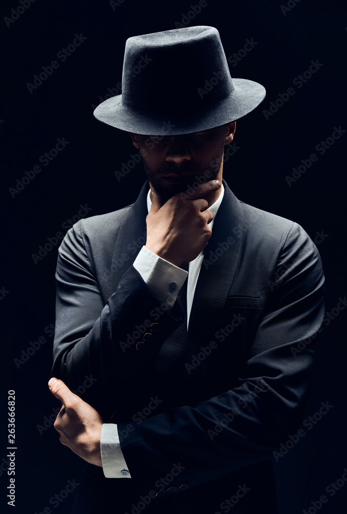 Handsome thoughtful man in black suit and hat touching chin on dark background Handsome thoughtful man in black suit and hat touching chin on dark background