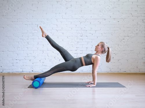 Caucasian young woman in grey sports suit doing pilates workout on math with fitness roller, loft style background, toned light blue