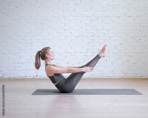 Beautiful girl in grey sports suit doing workout on math, boat classic yoga pose, loft style background, toned light blue