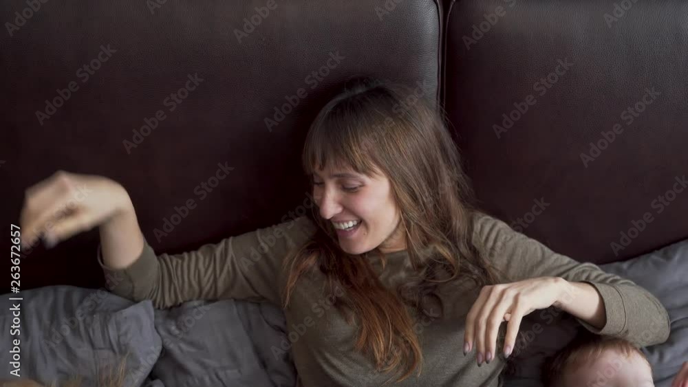 Happy mother with her three children having fun all together while wearing pijamas, on a big modern bed, smiling and laughing. Mother stretching while kids laughing and playing Happy mother with her three children having fun all together while wearing pijamas, on a big modern bed, smiling and laughing. Mother stretching while kids laughing and playing