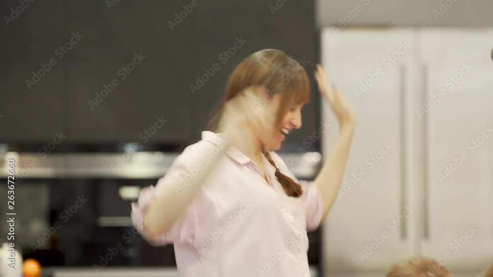 Happy mother having fun, clapping and smiling, with her kids while they are sitting on a table, while wearing pijamas, in a big and modern kitchen, with cooking equipment on the background Happy mother having fun, clapping and smiling, with her kids while they are sitting on a table, while wearing pijamas, in a big and modern kitchen, with cooking equipment on the background