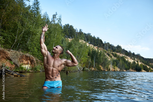 A man with perfect muscular arms, biceps, torso and abs stands in a classic bodybuilder pose outside in the nature