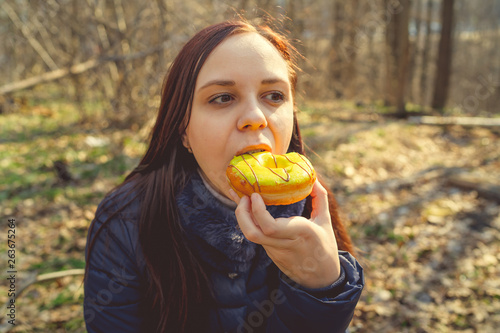 Young woman eating doughnut in woods Casual young brunette in jacket sitting on log in woods and eating delicious glazed doughnut